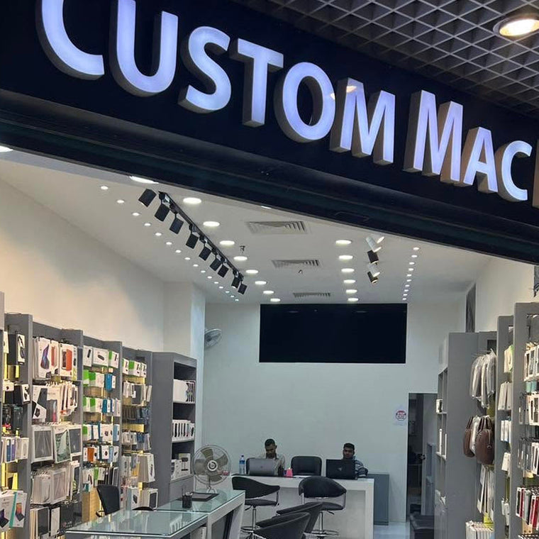 A retail storefront with a large illuminated sign reading 'CUSTOM MAC BD' above a modern interior. The shop has gray display walls, a glass-topped counter, white tiled floors, and several people seated or standing inside.